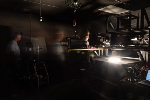 A long exposure image shows a man in a blue shirt walking across a dark room towards a table with a light in the middle.