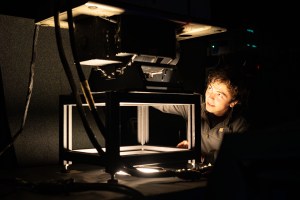 A boy with short black hair leans towards an instrument mounted on a table with a hole cut out of it and light coming up, illuminating his face. The rest of the room is dark.