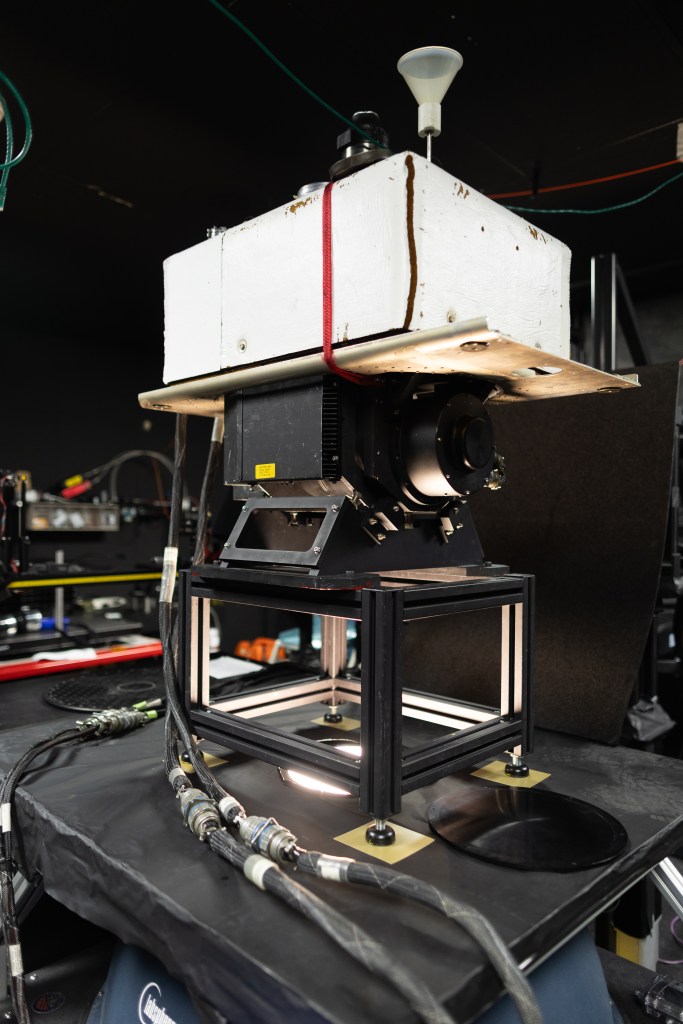 A large white box sits on top of a black instrument, on top of metal lets, all of which sit on a table with a hole cut out and light coming from within. The room is painted black.