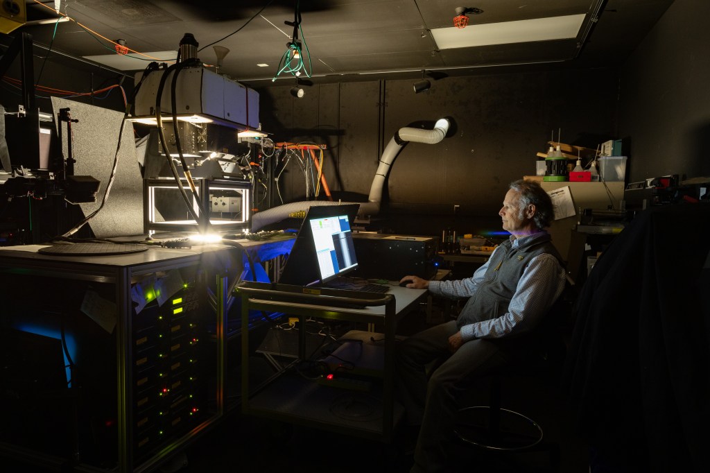A man with gray hair, a blue longsleeve and a gray vest sits at a desk looking at a monitor in a room painted all black. In the rest of the room are an assortment of machines and wires. The only light in the room is coming from hole cut out of a table in front of him.