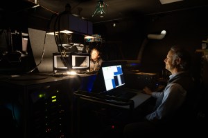 A man with gray hair, a blue longsleeve and a gray vest sits at a desk looking at a monitor in a dark room. The only light in the room is coming from hole cut out of a table in front of him, where another man looks up at the instrument on the table.