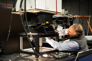 A man with gray hair, a blue longsleeve and a gray vest works on an instrument mounted on a table, over a hole with light coming out.