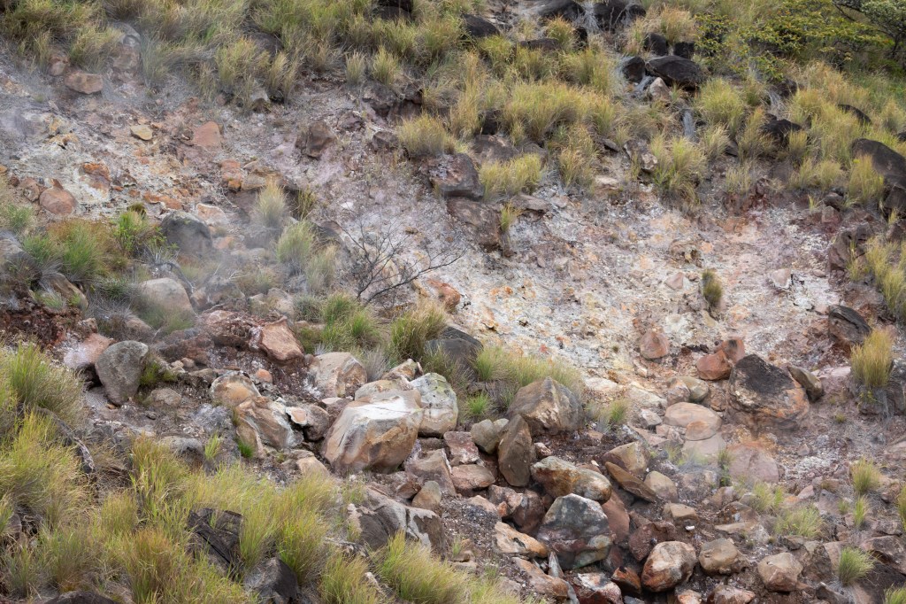 Reddish brown rocks sit on a slope with tufts of green grass. A bit of haze rises from the rocks.