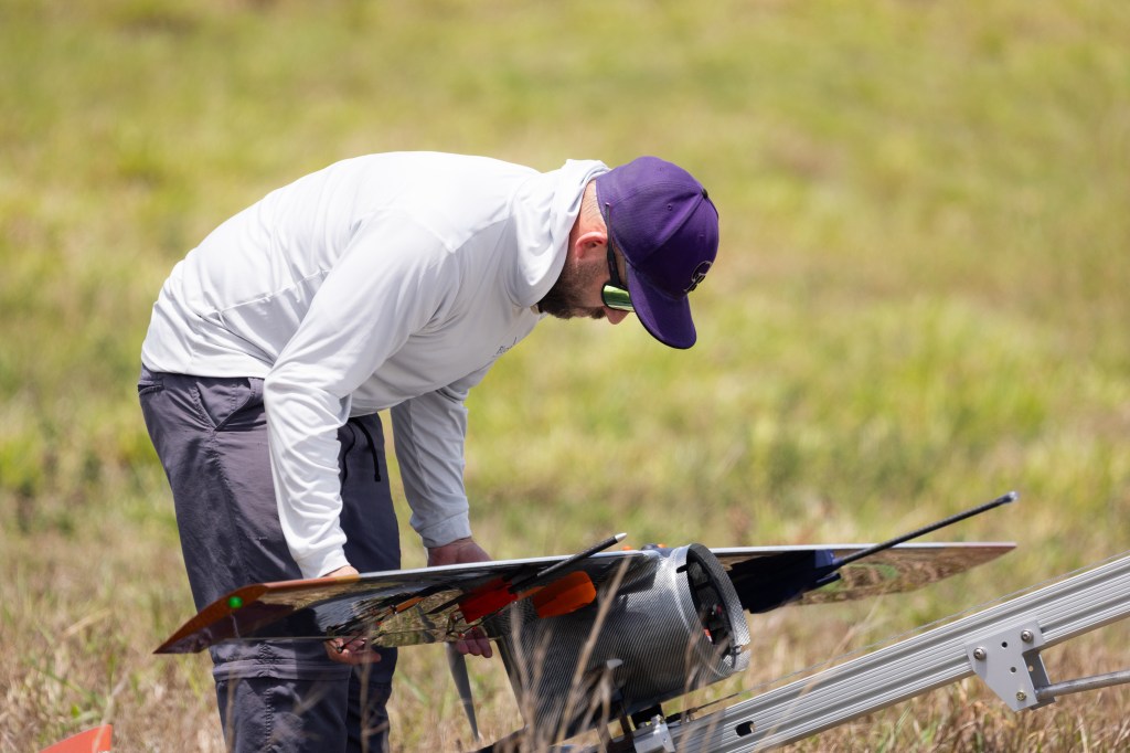 A man in a white longsleeve and purple baseball cap adjusts the wings on a slate-gray device.