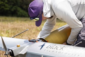 A man in a white longsleeve and purple baseball cap works on a gray aircraft that says Black Swift Technologies on the wings, next to a silhouette of a black bird.