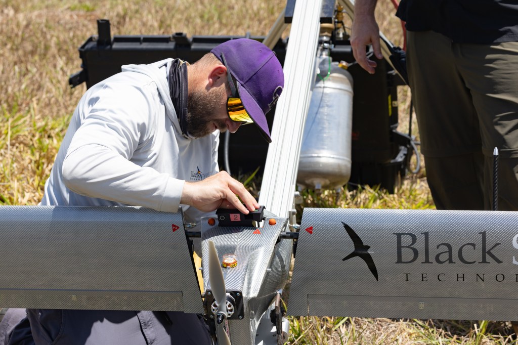 A man in a white longsleeve and purple baseball cap works on a gray aircraft that says Black Swift Technologies on the wings, next to a silhouette of a black bird.