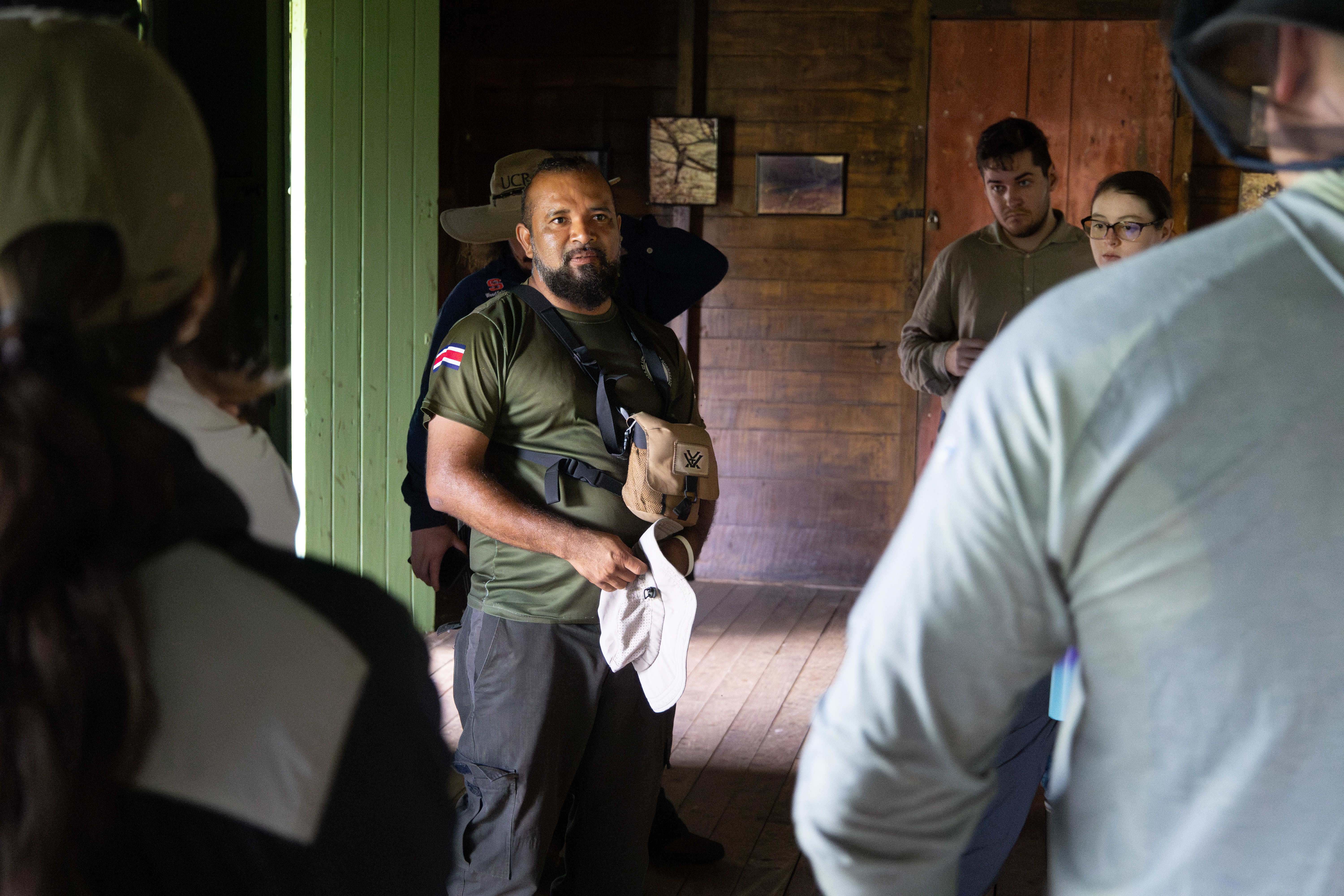 A man with a black beard, wearing an olive green t-shirt and black pants stands in a dark room speaking to a group of people.