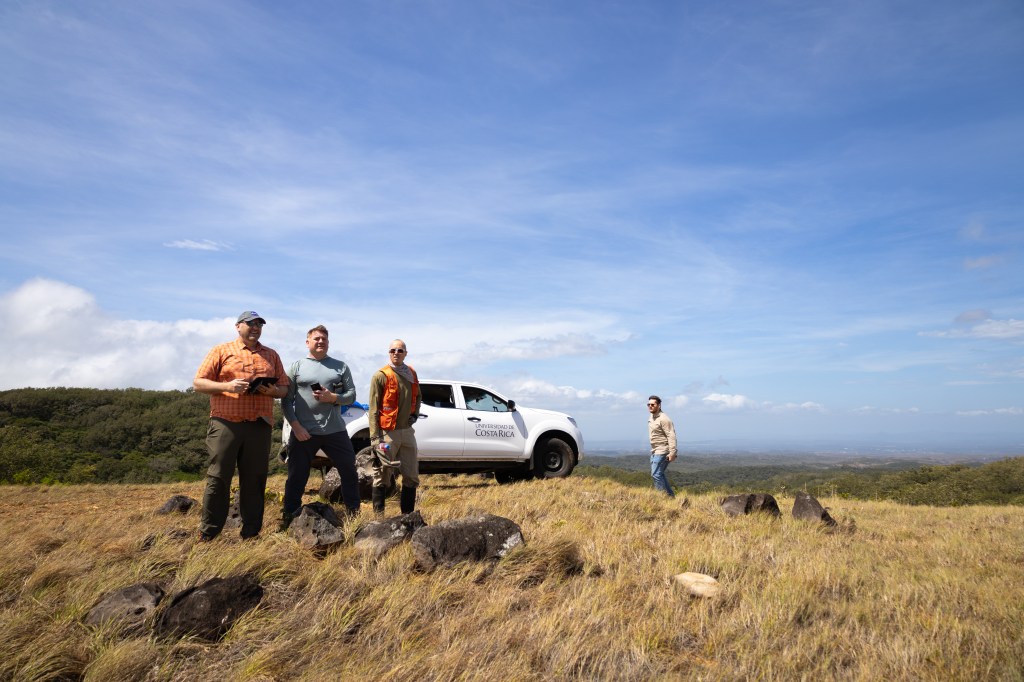 Three men stand on top of a hill of tan grass. In the background is a white pickup trust, a man walking along the ridge, a green forested hill, and a blue sky with wisps of white clouds.