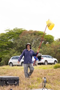 A man in a dark purple long sleeve shirt, gray hiking pants, and a white hat smiles at the camera while holding a long red pole with a yellow grocery bag tied to the end. In the background two white cars are parked on a grassy field.