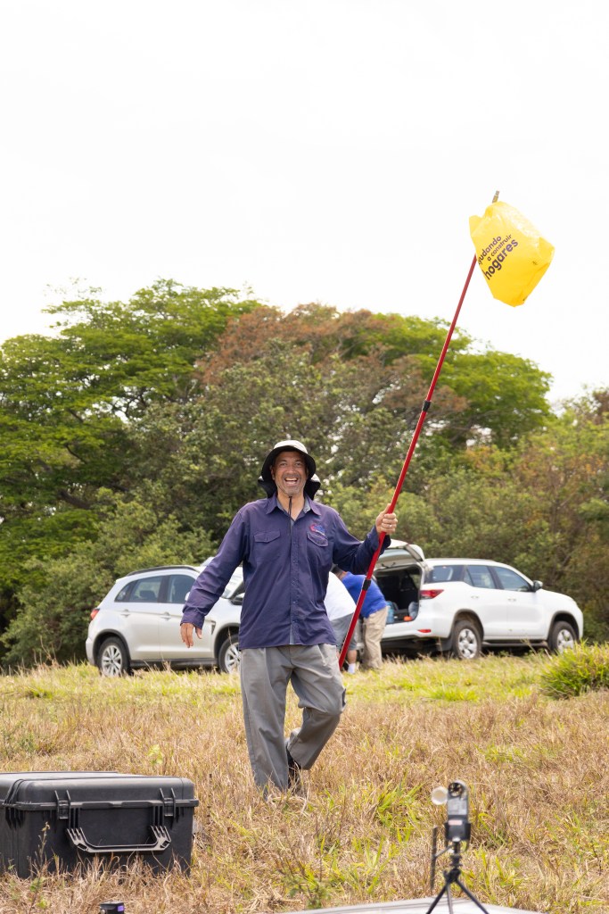 A man in a dark purple long sleeve shirt, gray hiking pants, and a white hat smiles at the camera while holding a long red pole with a yellow grocery bag tied to the end. In the background two white cars are parked on a grassy field.