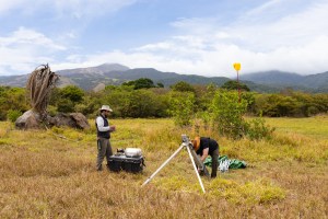 Two men work on something shaped like a silver tripod, with a small black pelican case, all in a green and tan field. In the distance is a green forested mountain and cloudy blue skies.