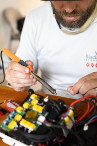 A close-up image of a man's hands holding a small soldering tool, positioned over a small circuit board.