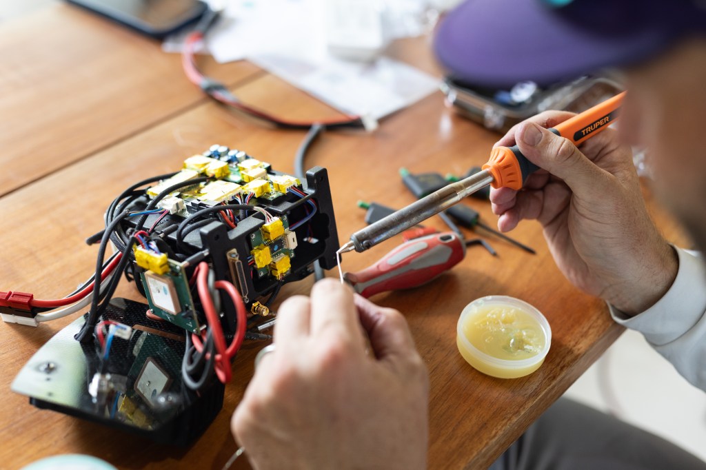 A close-up image of a man's hands holding a small soldering tool, positioned over a small circuit board.