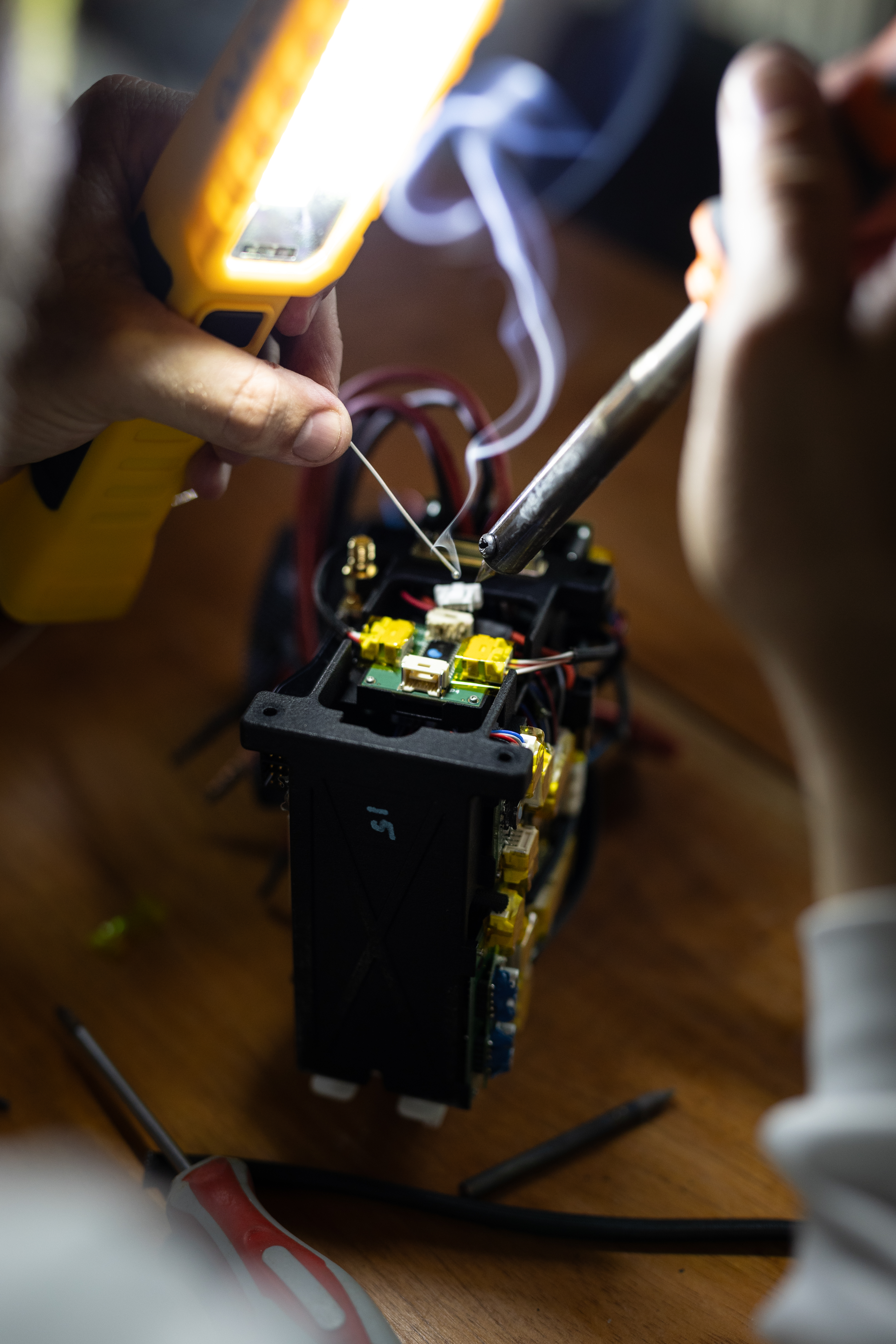 A close-up image of a man's hands holding a small soldering tool, positioned over a small circuit board.