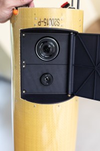 A black flap is open on a yellowish cylinder, revealing two circular lenses embedded on a black panel.