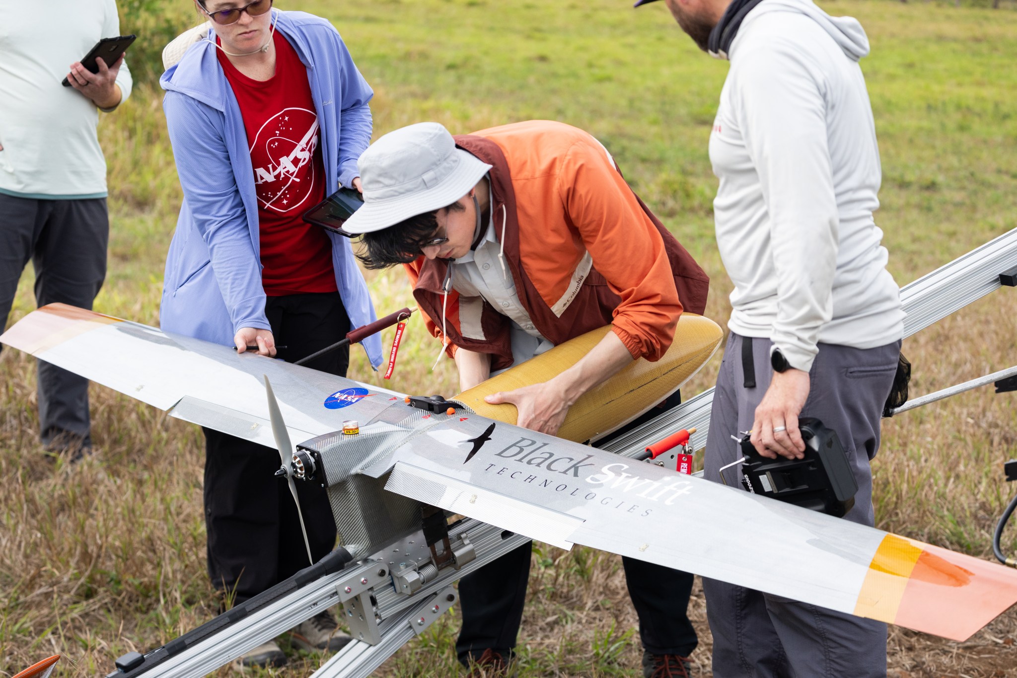 A man with black hair, a white bucket hat and an orange jacket bends over a small gray aircraft to attach a yellow nose cone.