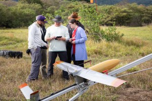 In the foreground is a small uncrewed aircraft with light gray wings and a yellow nose cone. In the background a group of four people in outdoorsy clothing gather around an ipad. The ground is covered in tan grass, which turns into green trees in the back and blueish hills in the distance.