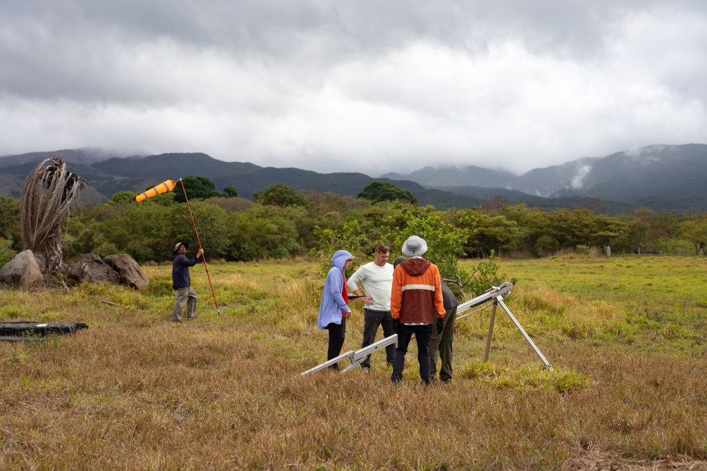 Three people in (from left) blue, white, and orange long sleeves stand next to a silver launcher shaped roughly like a large tripod. In the back, a man in black carries a long red pole with an orange windsock on the end. The ground is covered in red and yellow grass, which turns into green trees in the background and blueish mountains behind, under cloudy skies.