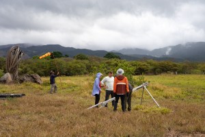 Three people in (from left) blue, white, and orange long sleeves stand next to a silver launcher shaped roughly like a large tripod. In the back, a man in black carries a long red pole with an orange windsock on the end. The ground is covered in red and yellow grass, which turns into green trees in the background and blueish mountains behind, under cloudy skies.