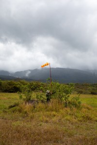 A man in black, wearing a white bucket hat, places a long red pole with an orange windsock into a green shrub. The ground is covered in red and yellow grass, which turns into green trees in the background and blueish mountains behind, under cloudy skies.