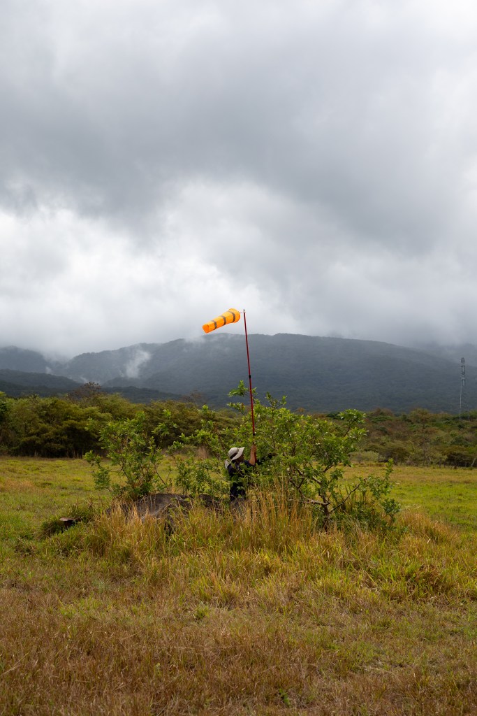 A man in black, wearing a white bucket hat, places a long red pole with an orange windsock into a green shrub. The ground is covered in red and yellow grass, which turns into green trees in the background and blueish mountains behind, under cloudy skies.