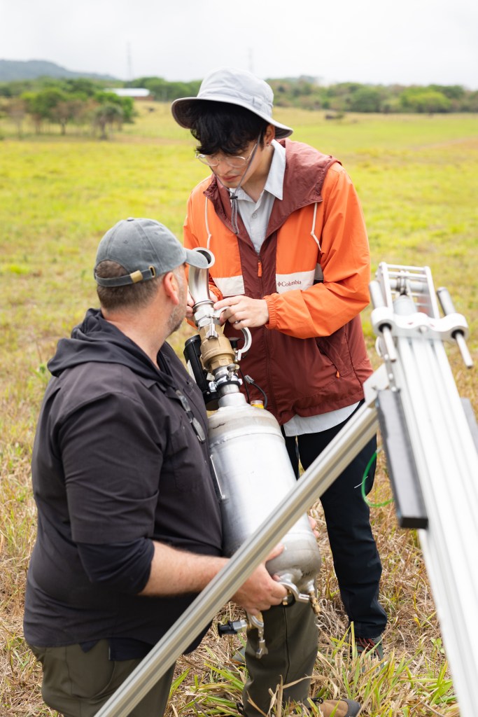 A man in black with a gray baseball cap kneels on the ground holding a large silver cylinder, while another man in an orange jacket and white bucket hat adjusts mechanics on the end of the cylinder.
