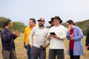 A group of six people stand in a field of tan grass, some speaking to each other and some looking out in the distance to the left of the frame.