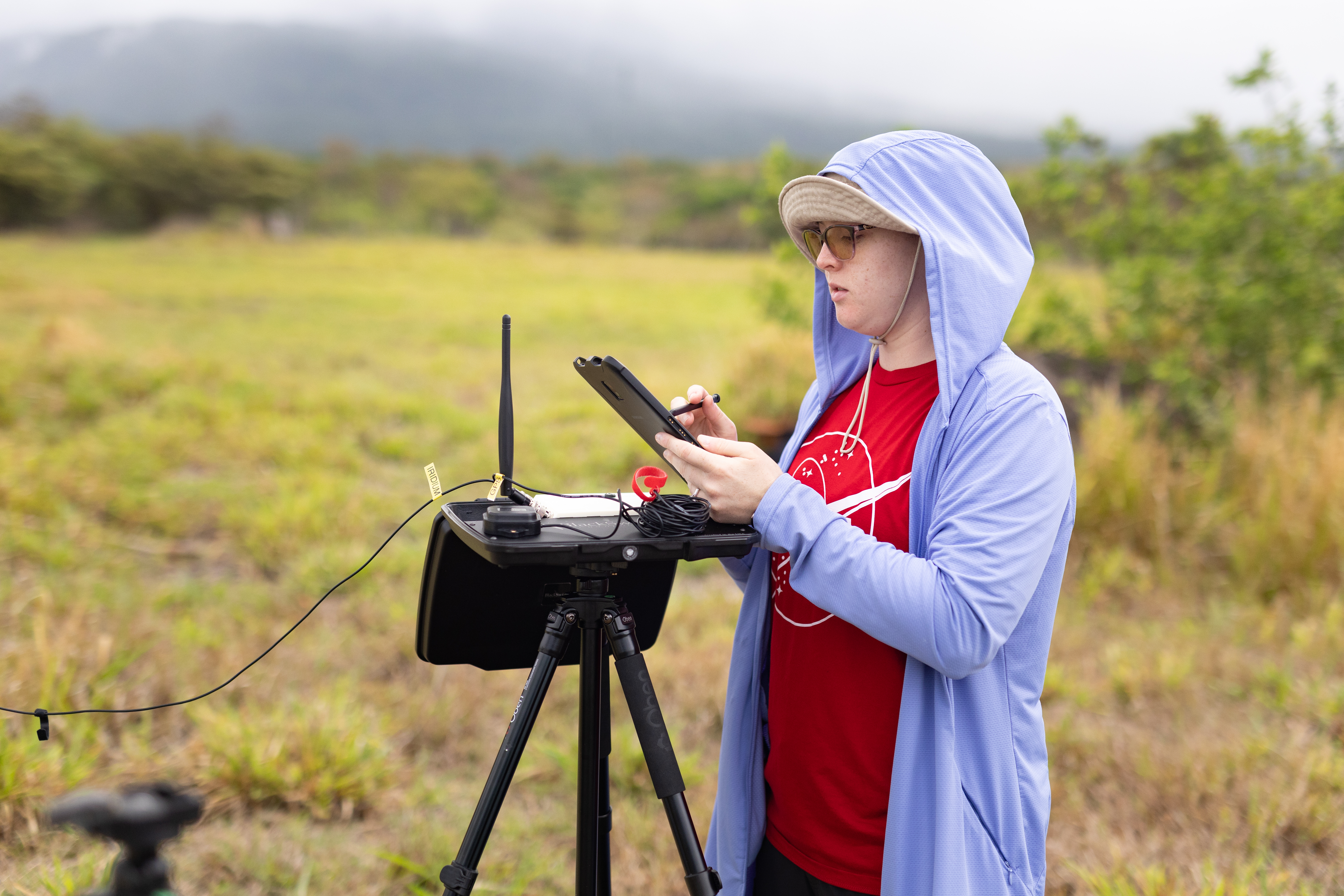 A woman in a red t-shirt and a light periwinkle jacket with the hood up looks at a tablet device. Next to her is a black tripod holding a black box with an antenna.