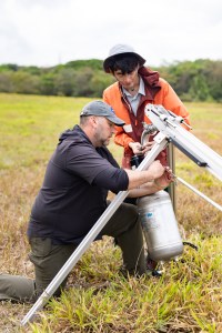 A man in black with a gray baseball cap kneels on the ground attaching a large silver cylinder to a silver tripod structure, while another man in an orange jacket and white bucket hat adjusts mechanics on the end of the cylinder.