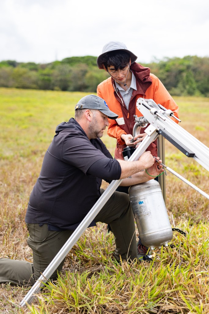 A man in black with a gray baseball cap kneels on the ground attaching a large silver cylinder to a silver tripod structure, while another man in an orange jacket and white bucket hat adjusts mechanics on the end of the cylinder.