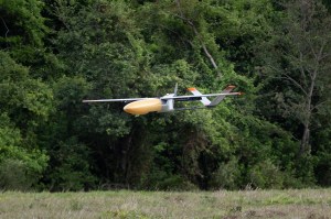 An gray uncrewed fixed-wing aircraft with a yellow nose cone flies towards the left of the frame against bright green foliage