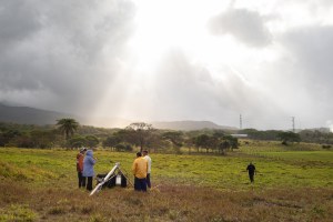 Four people stand around a silver tripod structure, in a red and green grassy field. Across the field, a man in all black walks back towards the group. In the distance are trees and mountains, with sun breaking through gray clouds.