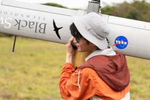 A man in an orange jacket and white bucket hat faces the left side of the frame, carrying a fixed-wing uncrewed aircraft over his shoulder. On one wing is the words Black Swift Technologies in a serif font with a silhouette of a black bird. On the other wing is the NASA logo: a blue circle with a red vector and the word NASA in white text.