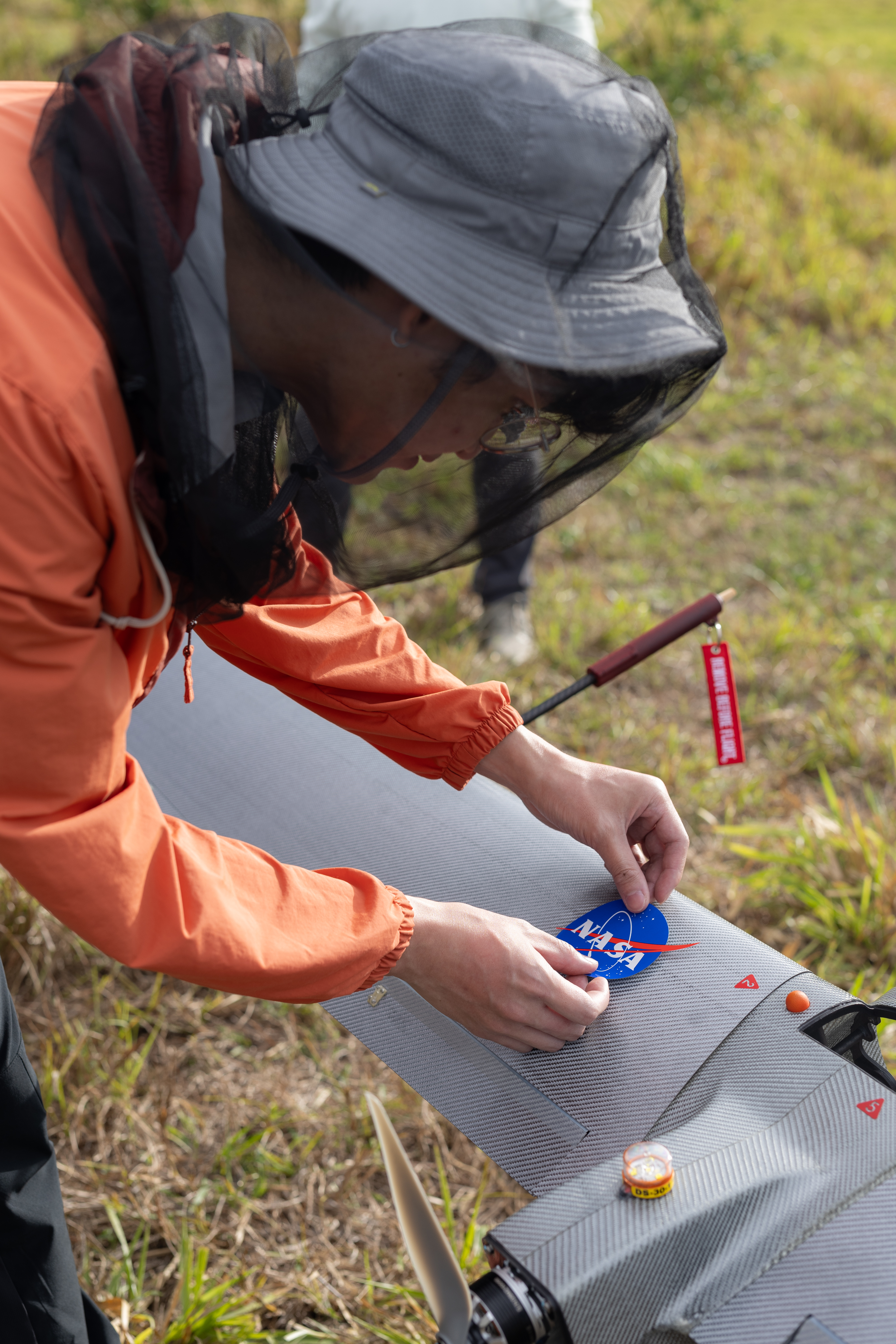 A man in an orange jacket, white bucket hat, and mosquito net leans down, placing a blue, circular NASA logo sticker onto a gray wing.