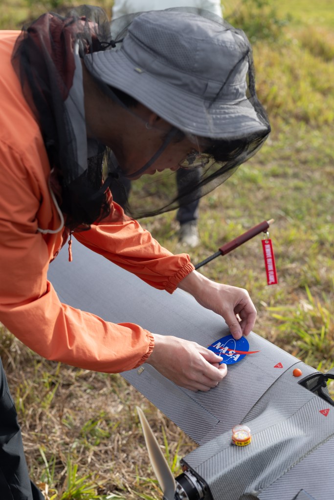 A man in an orange jacket, white bucket hat, and mosquito net leans down, placing a blue, circular NASA logo sticker onto a gray wing.