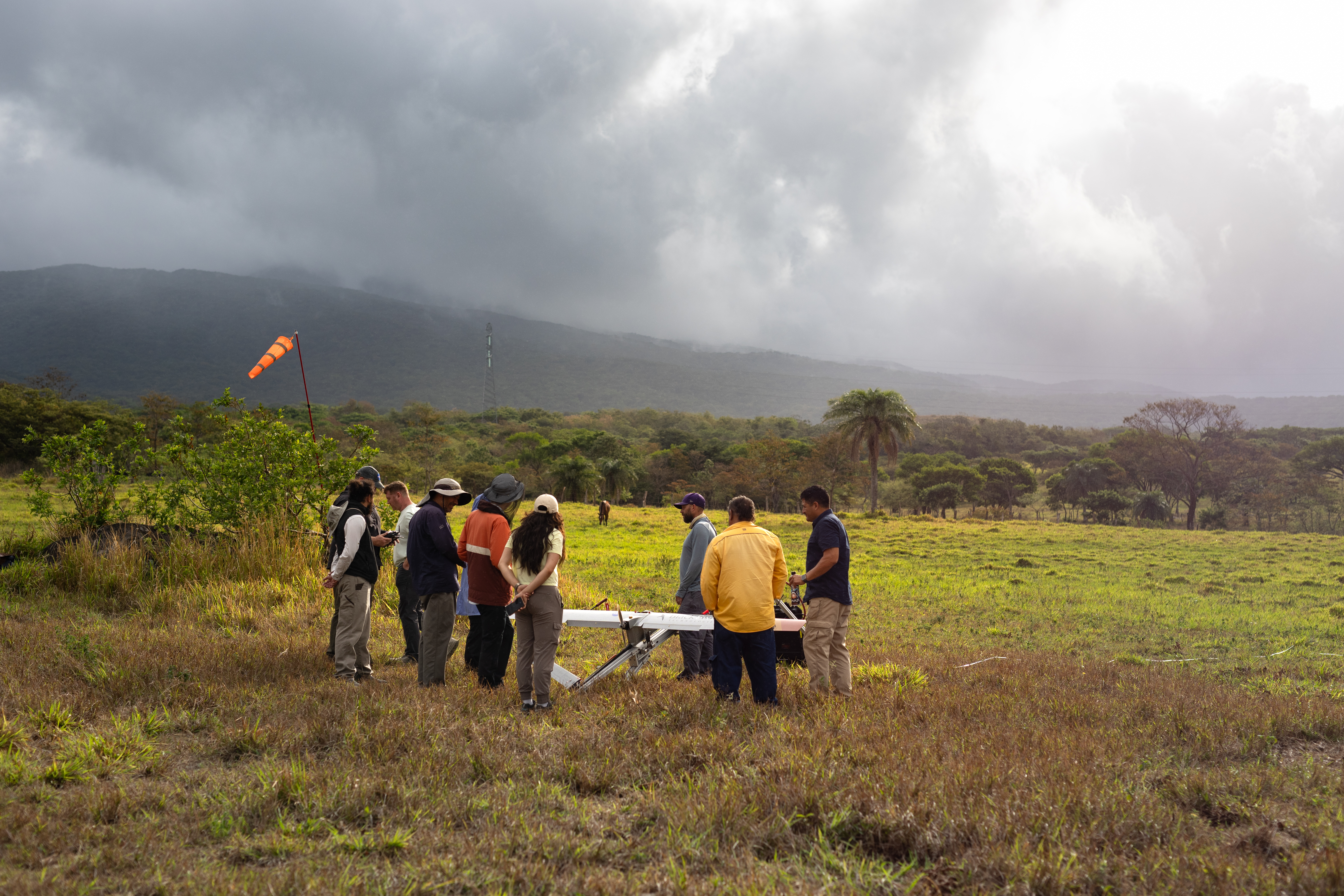 A group of people stand around a small gray fixed-wing aircraft mounted on a launcher, all in a grassy field. In the background are trees, a mountain, and stormy skies with the sun peeking through.