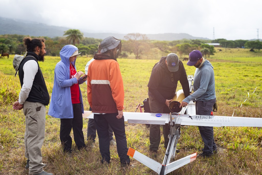 A group of five people stand around a small gray fixed-wing aircraft mounted on a launcher, all in a grassy green field.