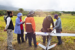 A group of five people stand around a small gray fixed-wing aircraft mounted on a launcher, all in a grassy green field.