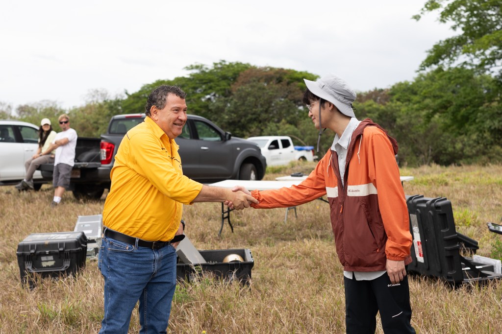 A man in a yellow long sleeve and jeans in the left of the frame shakes hands with a man in an orange jacket and white bucket hat in the right of the frame. In the background is a tan field littered with black boxes and pickup trucks.