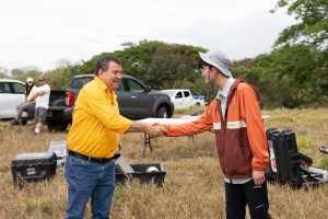 A man in a yellow long sleeve and jeans in the left of the frame shakes hands with a man in an orange jacket and white bucket hat in the right of the frame. In the background is a tan field littered with black boxes and pickup trucks.