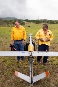 Two men in yellow long sleeve shirts and jeans laugh and look off to the left of the photo, posing behind a light gray fixed-wing aircraft with a yellow nose cone facing away from the camera.