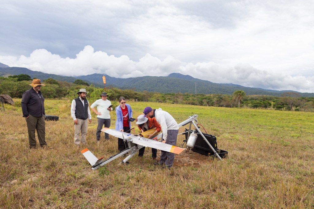 A group of six people stand in a field of tan and green grass, some looking at and some working on a small gray fixed-wing aircraft with a yellow nose cone, sitting on a silver launcher. In the background is a blueish mountain and cloudy skies.