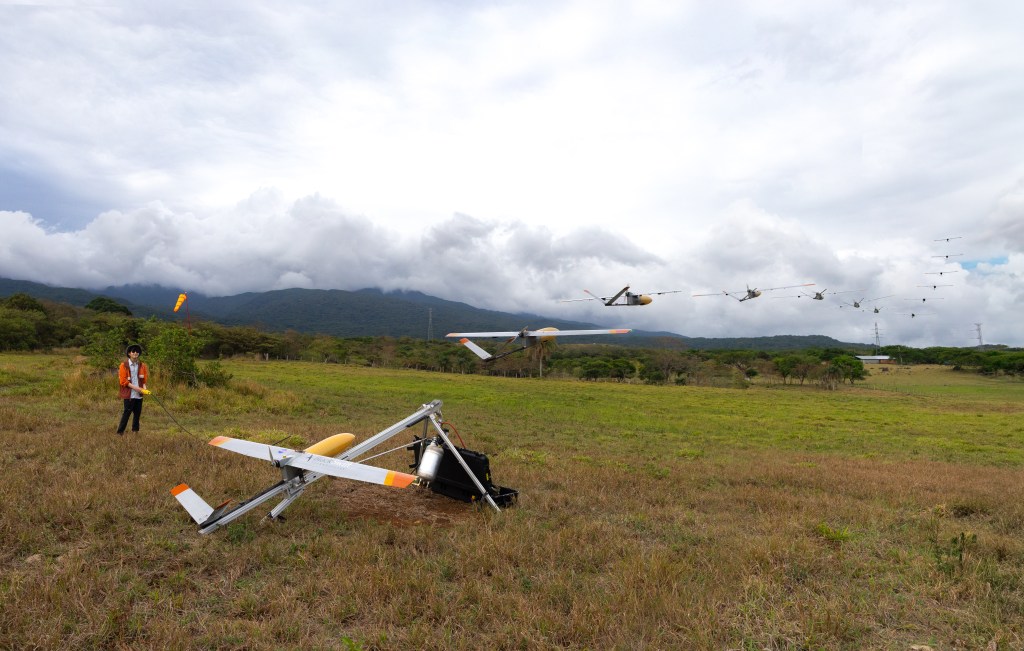 A man in an orange jacket and black pants stands in a field of tan and green grass, looking at a small gray fixed-wing aircraft with a yellow nose cone, sitting on a silver launcher. In this composite image, the aircraft is pictured again and again across the sky, moving away from the camera. In the background is a blueish mountain and cloudy skies.