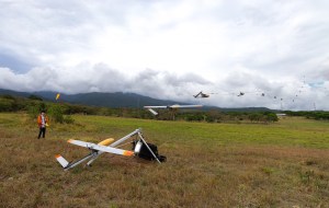 A man in an orange jacket and black pants stands in a field of tan and green grass, looking at a small gray fixed-wing aircraft with a yellow nose cone, sitting on a silver launcher. In this composite image, the aircraft is pictured again and again across the sky, moving away from the camera. In the background is a blueish mountain and cloudy skies.
