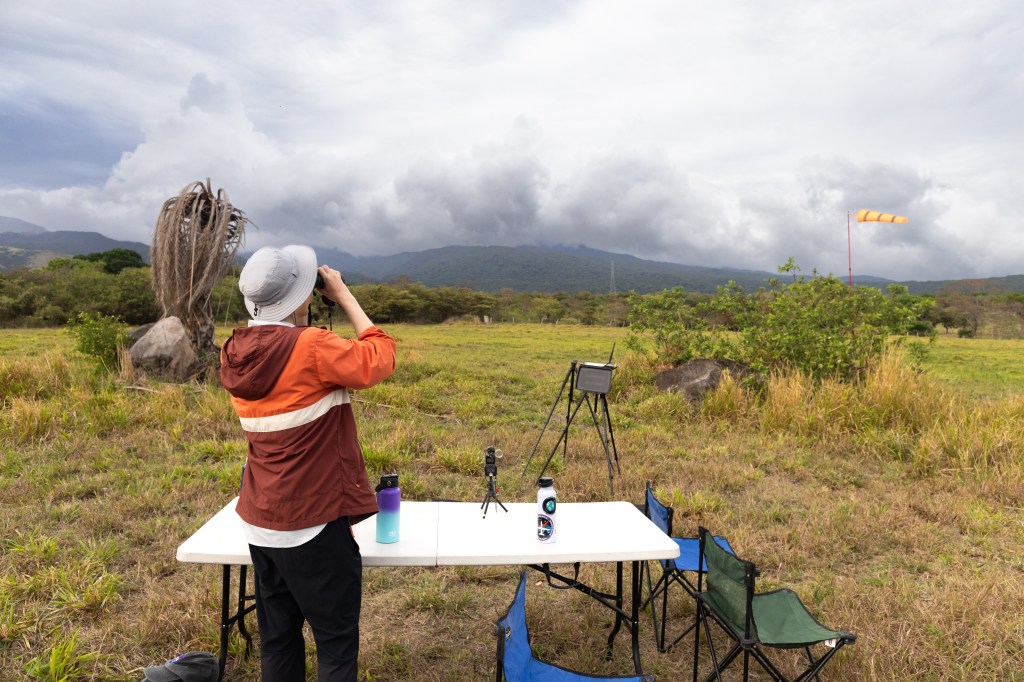 A man with an orange jacket, black pants, and white bucket hat stands at a white folding table with his back to the camera, using binoculars to look into the distance. He stands in a field, looking towards blueish mountains under a cloudy sky.