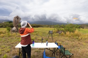 A man with an orange jacket, black pants, and white bucket hat stands at a white folding table with his back to the camera, using binoculars to look into the distance. He stands in a field, looking towards blueish mountains under a cloudy sky.