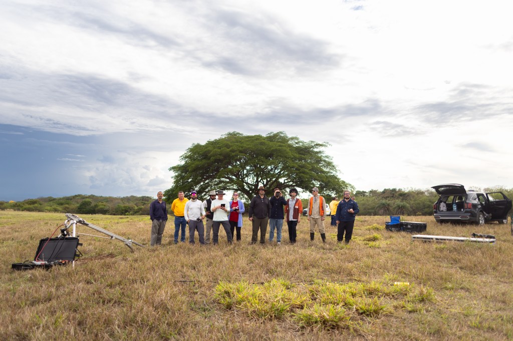A group of people stand in a line in a tan grassy field scattered with boxes and equipment, with a car parked in the back right and a large tree directly behind them. They all look in the same direction.