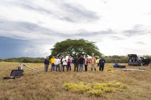 A group of people stand in a line in a tan grassy field scattered with boxes and equipment, with a car parked in the back right and a large tree directly behind them. They all look in the same direction.