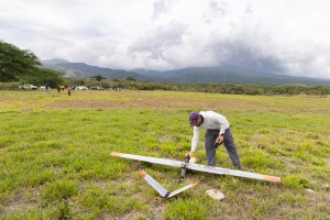 A man in a white long sleeve bends over a gray fixed-wing aircraft sitting in a bright green field, with blueish mountains and cloudy skies in the background.