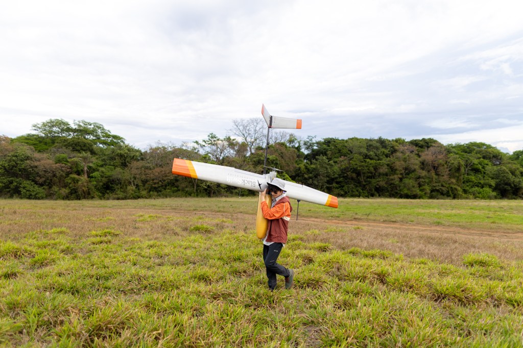 A man in an orange jacket, black pants, and white bucket hat faces the left side of the frame, carrying a fixed-wing uncrewed aircraft over his shoulder across a field of green grass. On one wing is the words Black Swift Technologies in a serif font with a silhouette of a black bird. On the other wing is the NASA logo: a blue circle with a red vector and the word NASA in white text.
